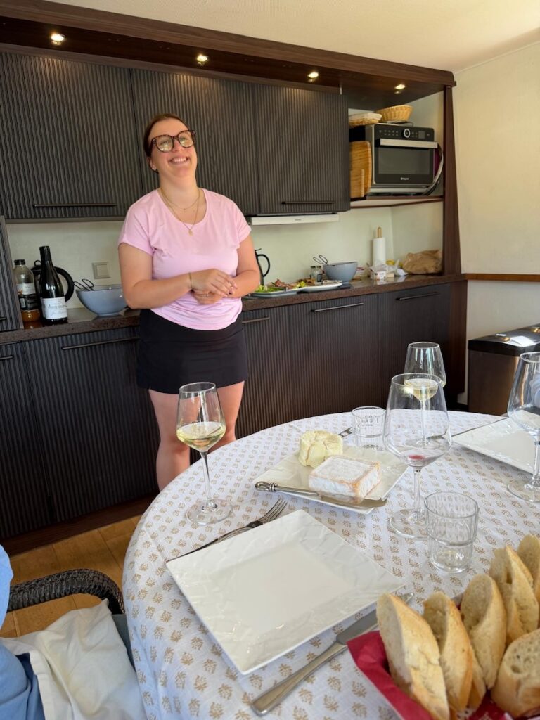 This French Barge Is Different (2026 REVIEW) — Choose The Savoir Vivre On The Burgundy Canal For The Cycling & Dinners Ashore 12 Matilda, the ship's hostesses stands in front of the wall of appliances that lines one side of the dining/living area on Savoir Vivre